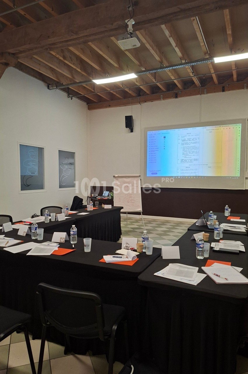 Salle de réunion avec tables disposées en U, documents, bouteilles d'eau et écran projetant un tableau coloré.