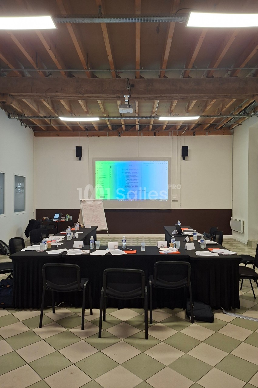 Salle de réunion avec tables disposées en U, écran de projection allumé, chaises et matériel de travail.