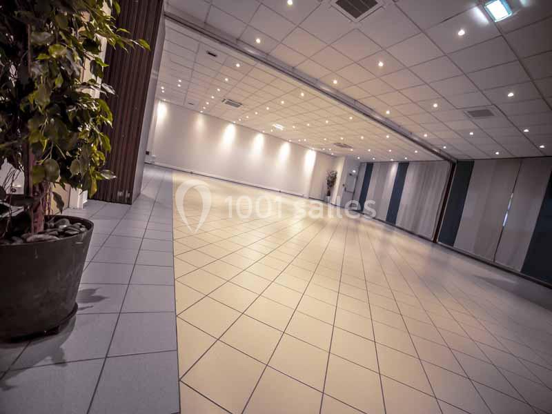 Salle vide avec carrelage clair, murs blancs, éclairage au plafond et une plante en pot au premier plan.