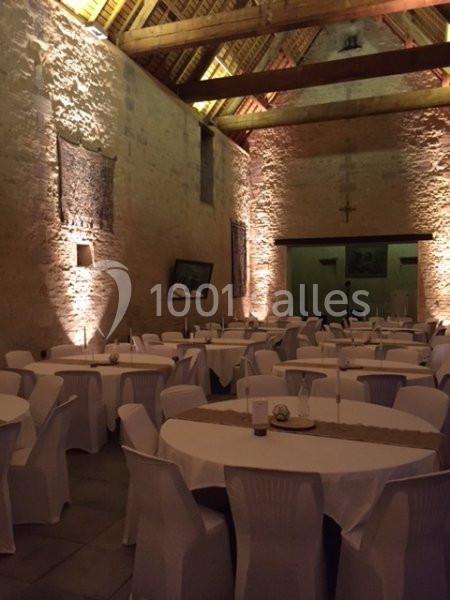 Salle en pierre avec des tables rondes dressées et des chaises blanches, éclairée par une lumière chaleureuse.