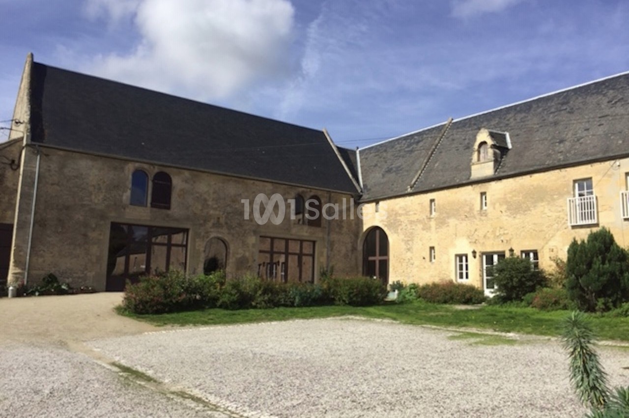 Bâtiments en pierre avec toits en ardoise entourant une cour gravillonnée sous un ciel partiellement nuageux.