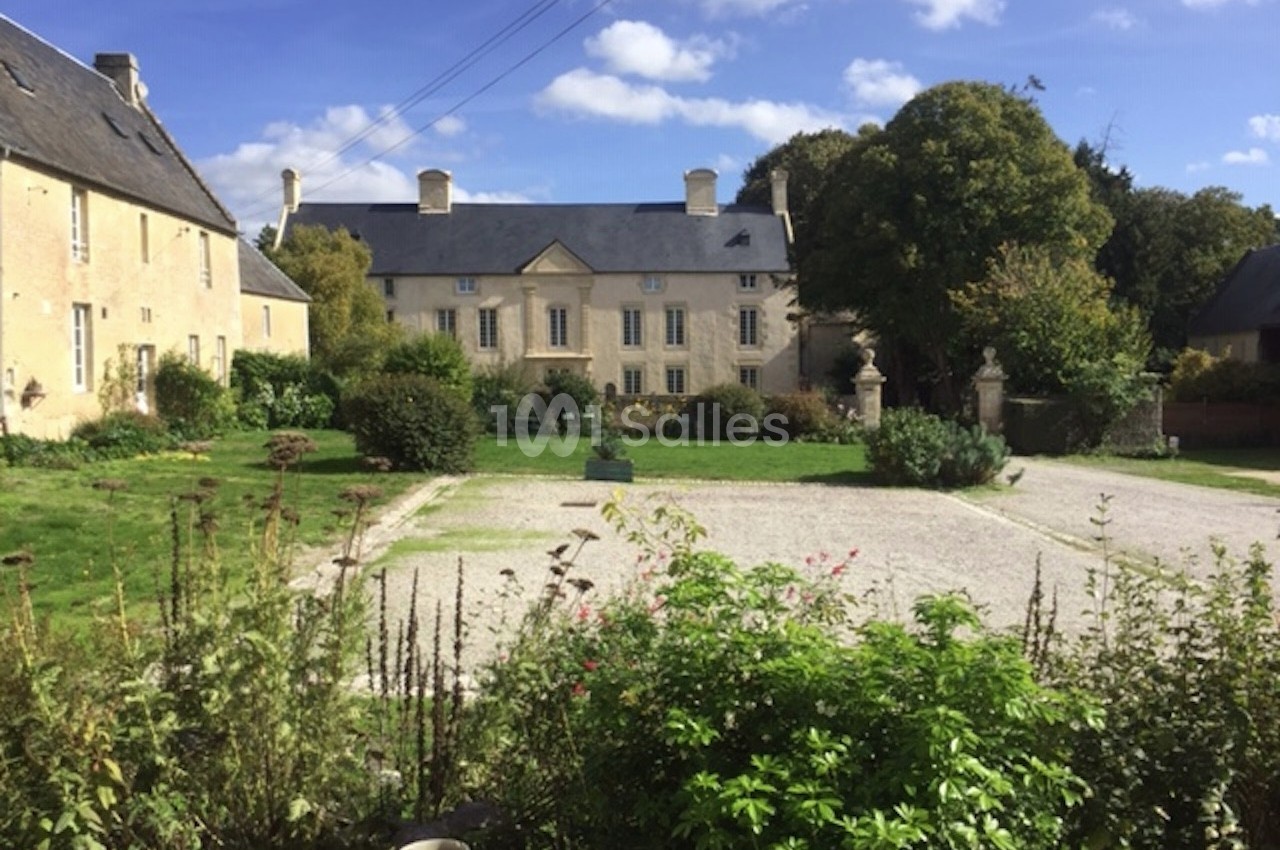 Cour d'une grande maison en pierre avec jardin, allée gravillonnée, arbres et ciel bleu parsemé de nuages.