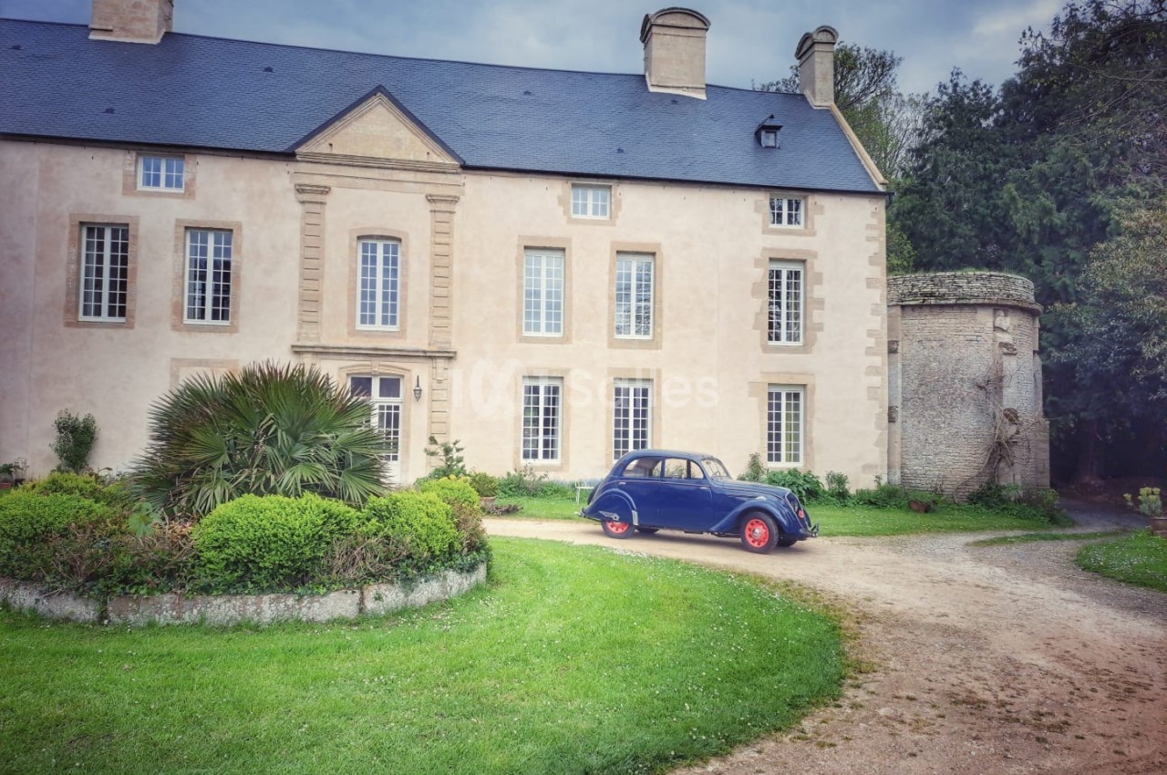 Façade d'une grande maison en pierre avec une voiture ancienne bleue garée devant, entourée de verdure.