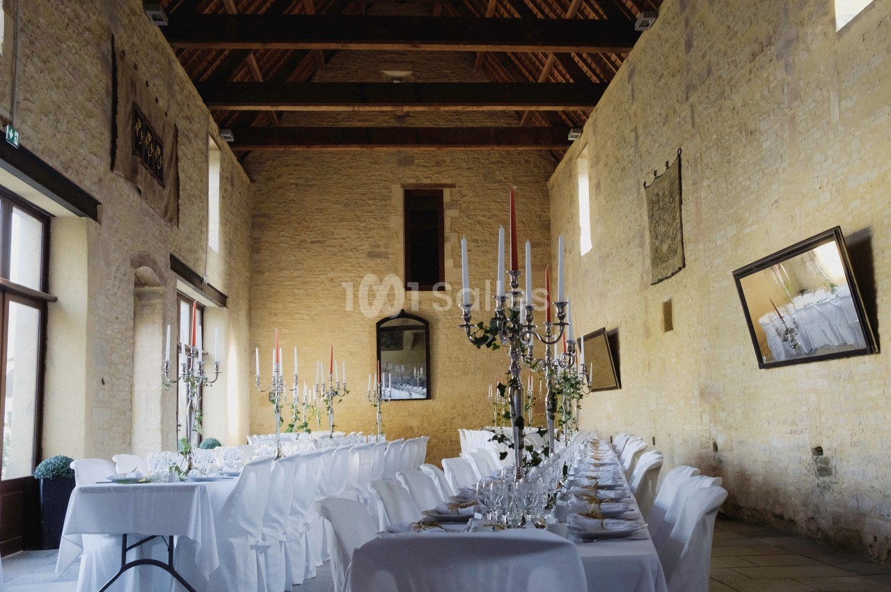 Grande salle en pierre avec tables dressées pour un banquet, ornées de chandeliers et de nappes blanches.