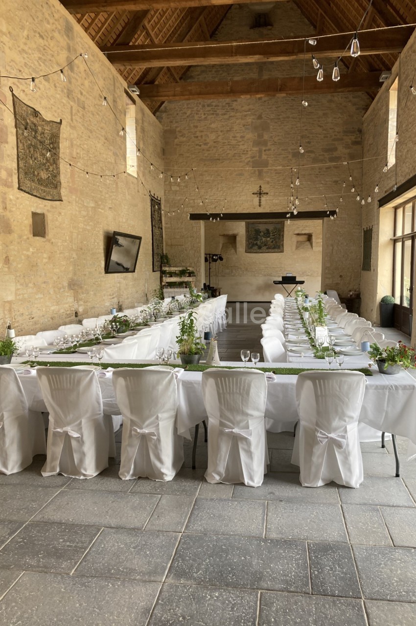 Salle en pierre avec tables dressées en U, nappes blanches, chaises couvertes, décorations végétales et guirlandes…