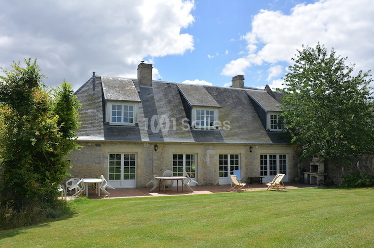 Maison en pierre avec toit en ardoise, grandes fenêtres, terrasse aménagée et jardin verdoyant sous un ciel partiellement…