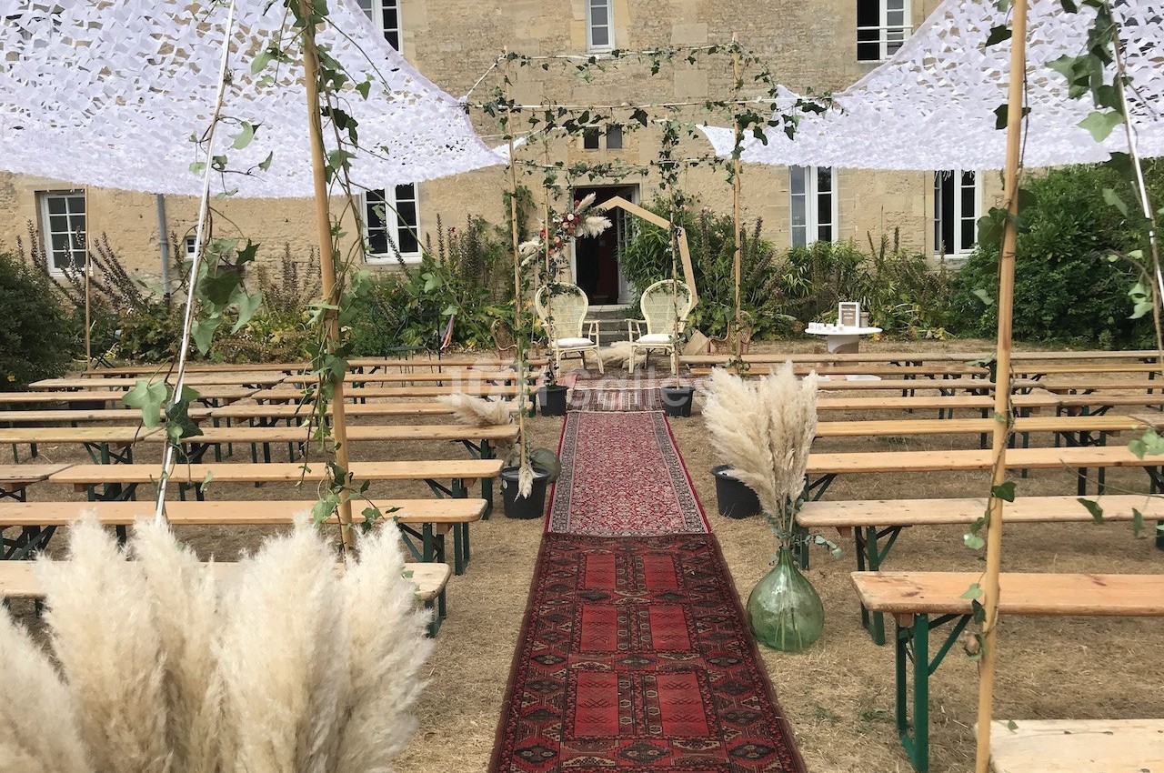 Allée centrale décorée de tapis rouges, entourée de bancs en bois, menant à une arche florale devant un bâtiment en pierre.