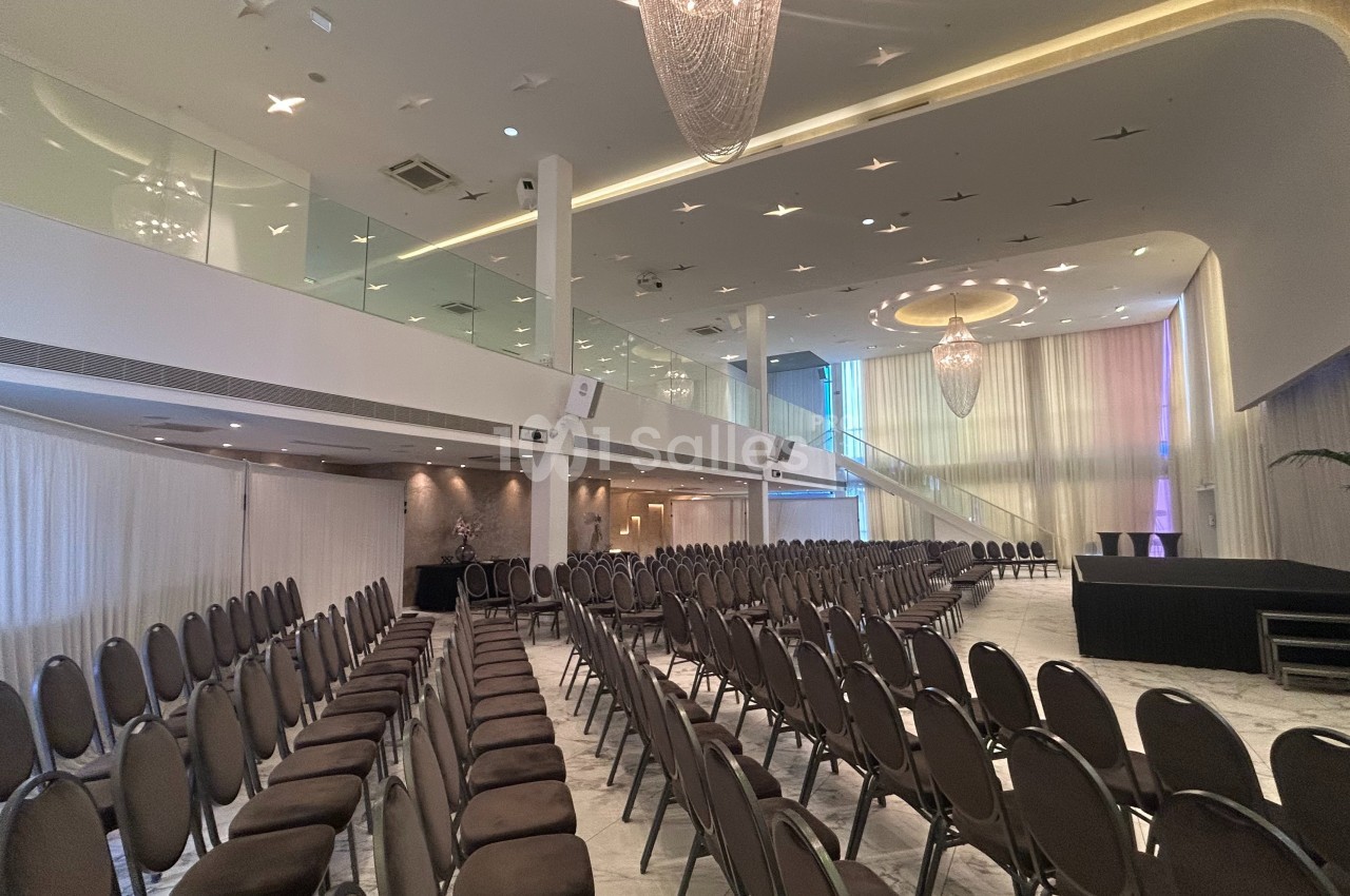 Salle de conférence moderne avec rangées de chaises alignées, scène surélevée et éclairage élégant.