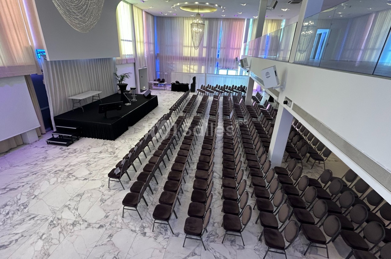 Salle de conférence lumineuse avec des rangées de chaises alignées face à une scène surélevée, sol en marbre.