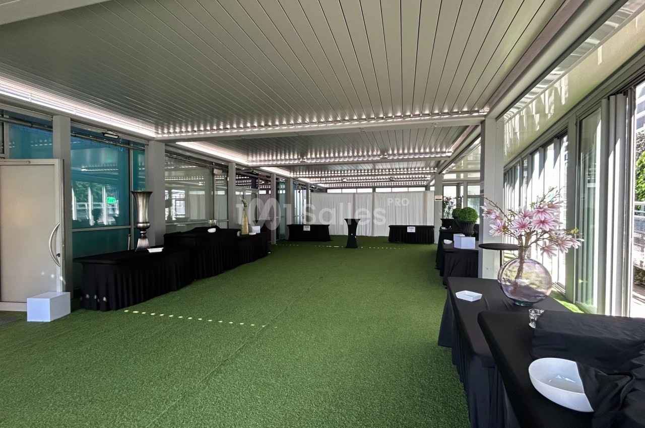 Salle lumineuse avec sol en gazon synthétique, tables noires alignées et décorations florales près de baies vitrées.