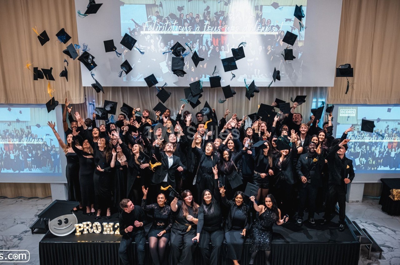 Des diplômés en tenue de cérémonie lancent leurs toques en l'air devant un écran affichant une photo de groupe.