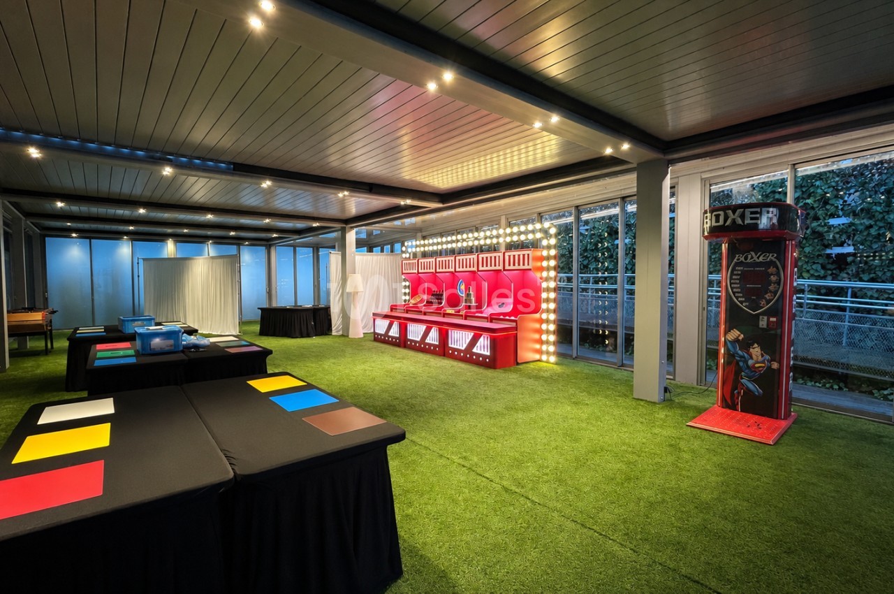 Salle de jeux intérieure avec stands colorés, machine de boxe et tables sur gazon synthétique, éclairage moderne.