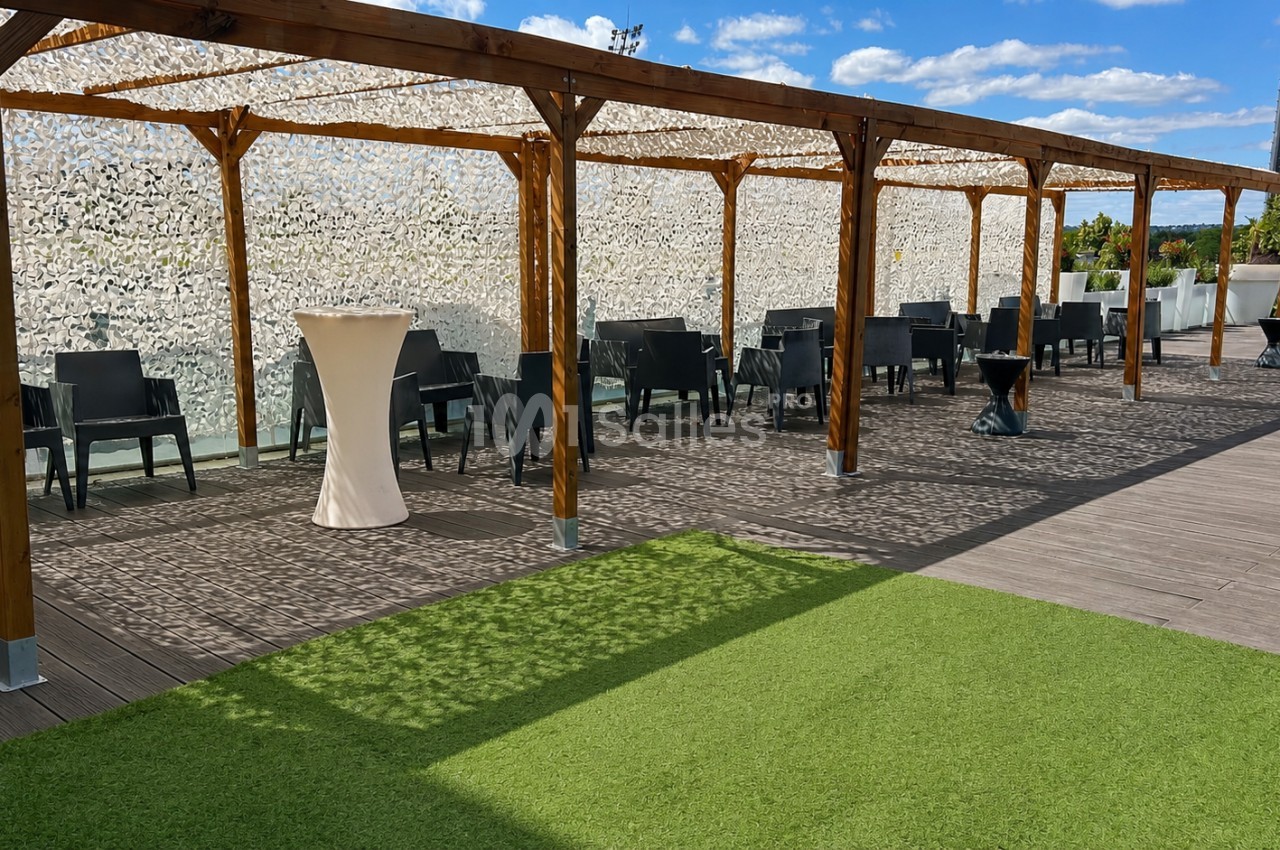 Terrasse avec pergola en bois, tables et chaises noires, sol en bois et gazon artificiel, ciel bleu en arrière-plan.