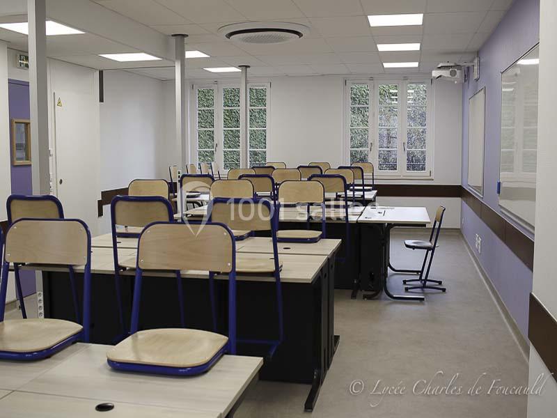 Salle de classe vide avec des bureaux en bois clair, des chaises empilées et des fenêtres donnant sur un espace arboré.