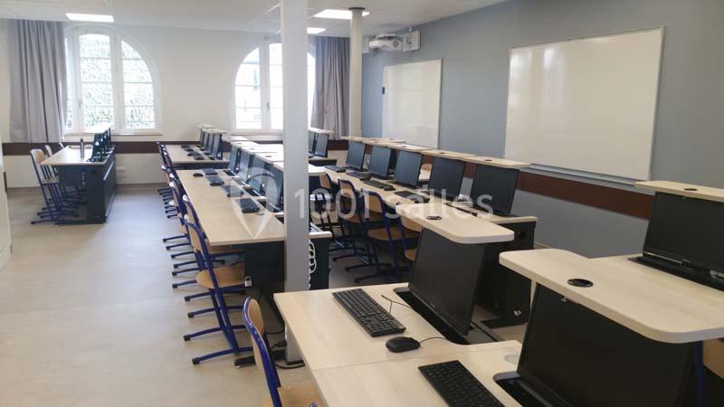 Salle informatique avec rangées de bureaux équipés d'ordinateurs, chaises et tableau blanc au fond.