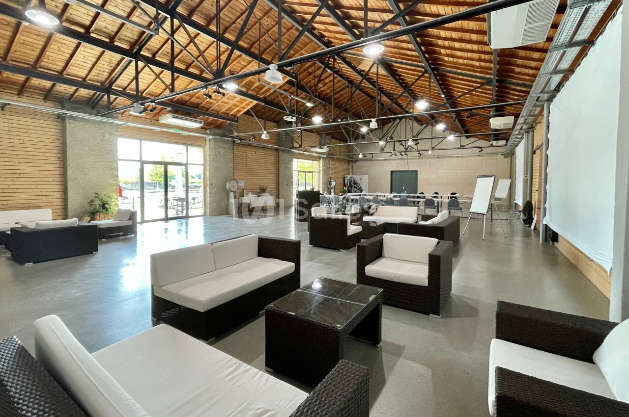 Salle spacieuse avec mobilier en rotin, grandes baies vitrées, plafond en bois et structure métallique apparente.