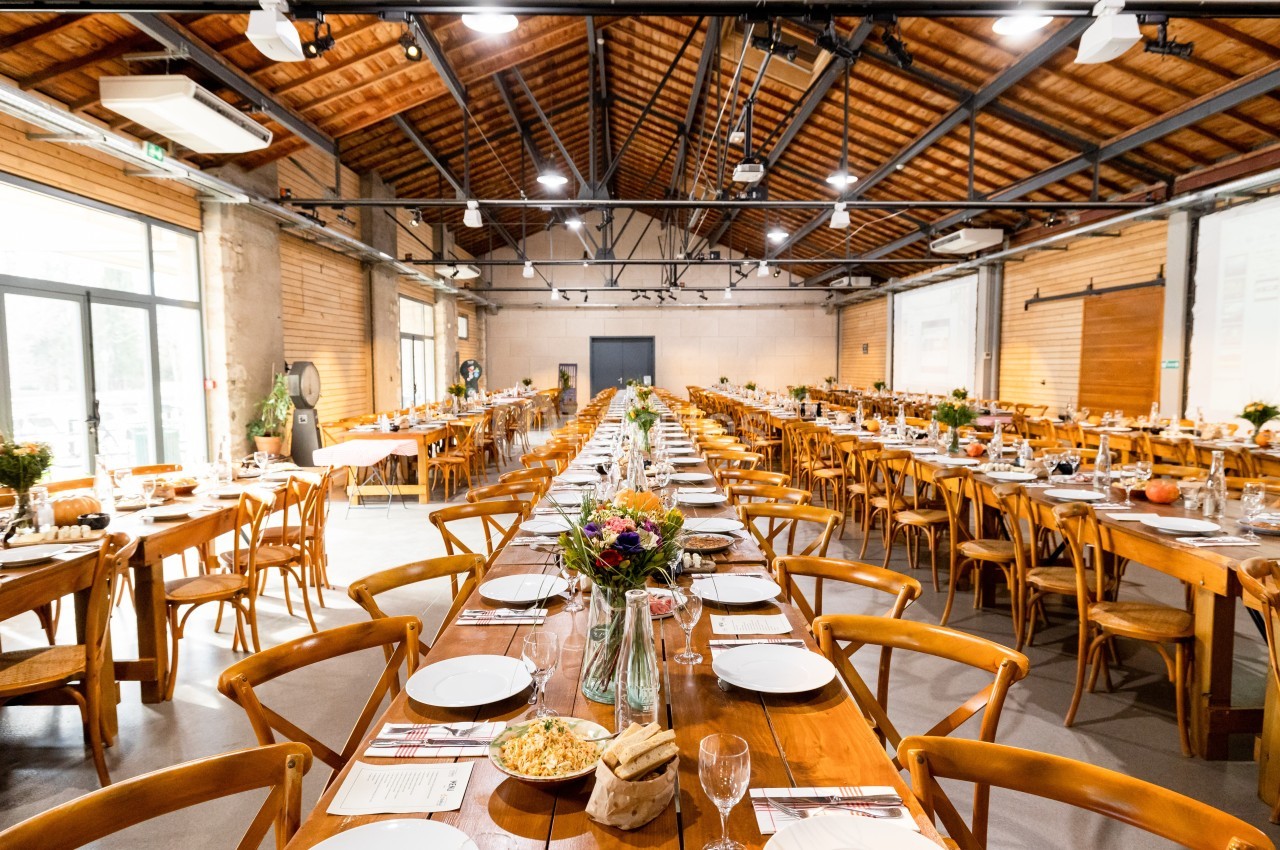 Salle de réception avec de longues tables en bois dressées pour un repas, décorée de fleurs et éclairée par des lumières…