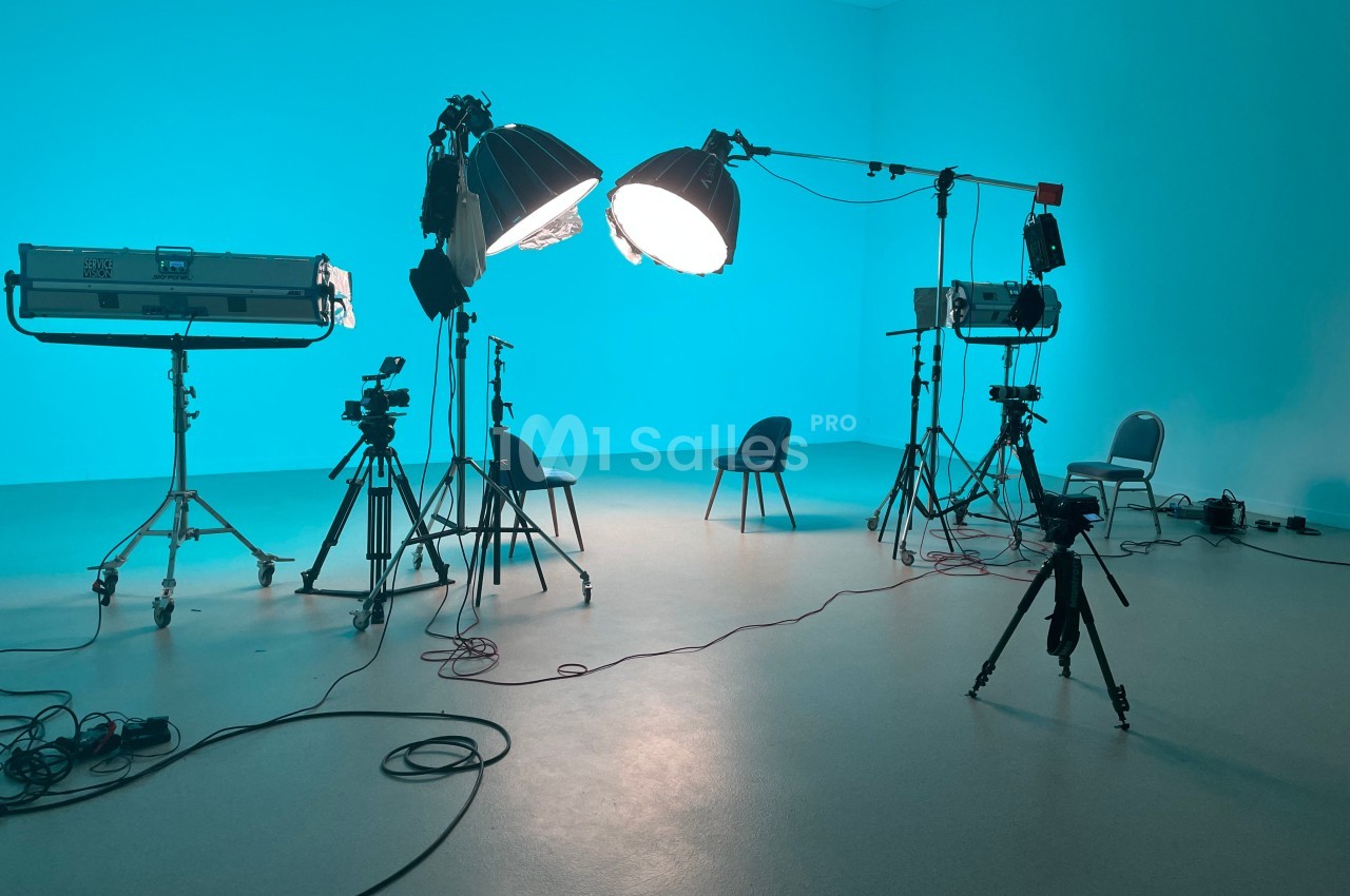 Studio photo avec éclairages, trépieds, caméras et chaises dans une pièce aux murs bleus.