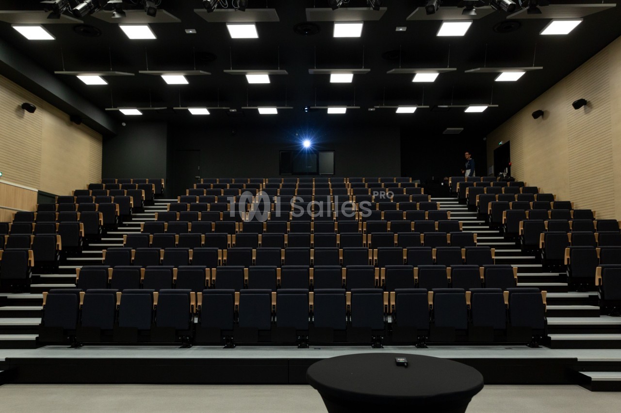Salle de conférence moderne avec rangées de sièges noirs vides et éclairage au plafond.
