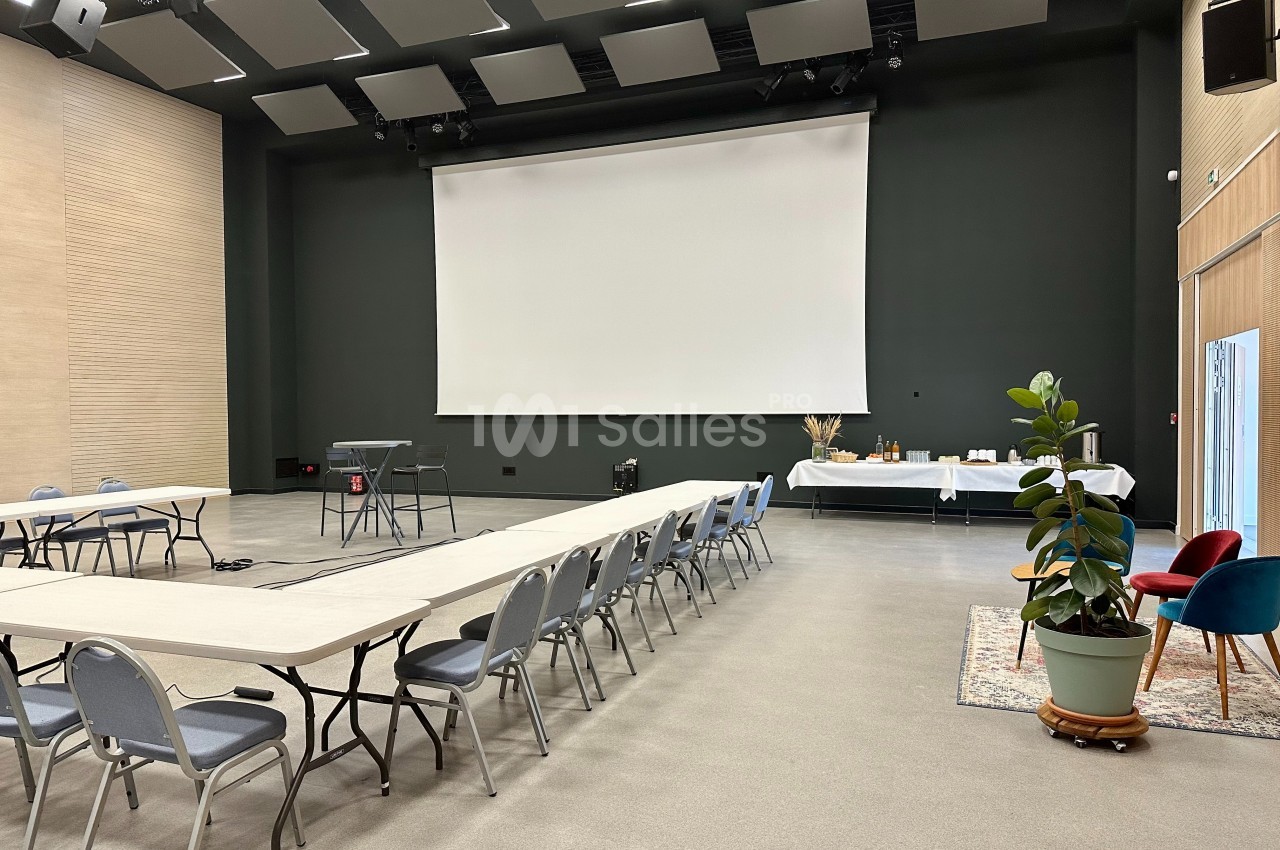 Salle de réunion avec tables et chaises disposées en U, grand écran blanc, plantes et buffet en arrière-plan.