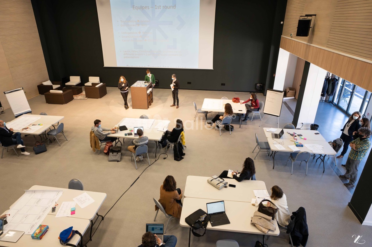 Salle de conférence avec des participants travaillant en groupes autour de tables et une présentation projetée à l'écran.