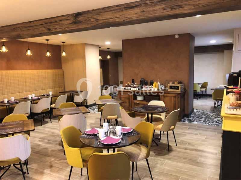 Salle de petit-déjeuner lumineuse avec tables dressées, chaises jaunes et buffet en bois proposant des boissons et…