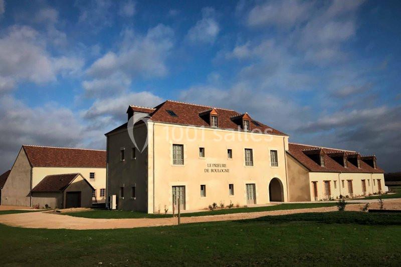 Bâtiment en pierre claire avec toit en tuiles rouges, entouré d'une pelouse et d'un ciel partiellement nuageux.
