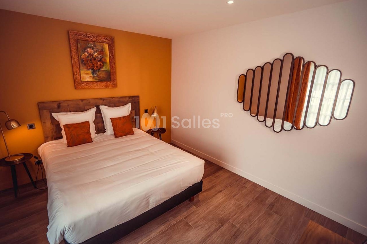 Chambre avec lit double, mur ocre, tableau floral, miroir design et sol en parquet.