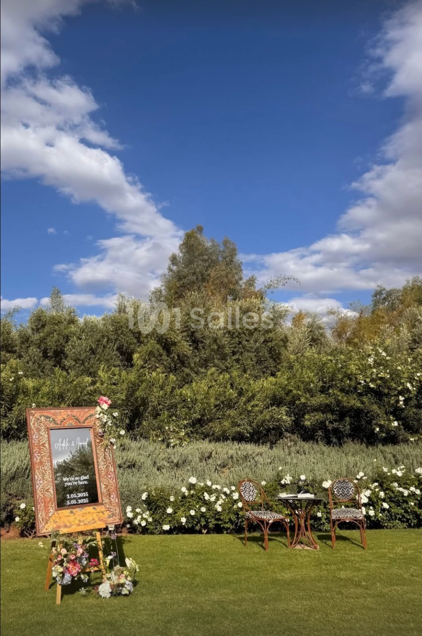 Panneau décoratif entouré de fleurs sur une pelouse, avec deux chaises et une table, sous un ciel bleu.