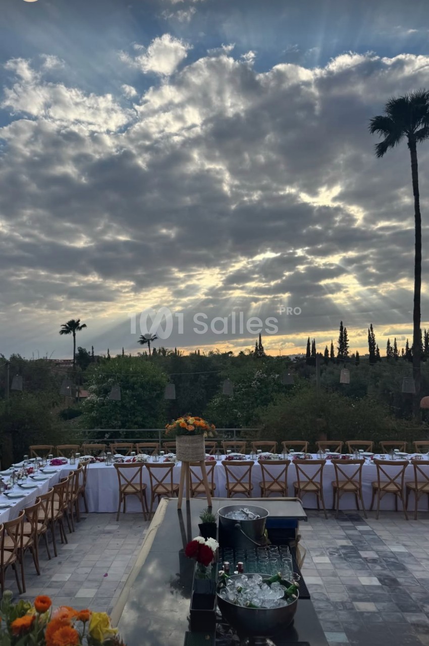 Terrasse extérieure avec tables dressées pour un repas, sous un ciel nuageux laissant passer des rayons de soleil.