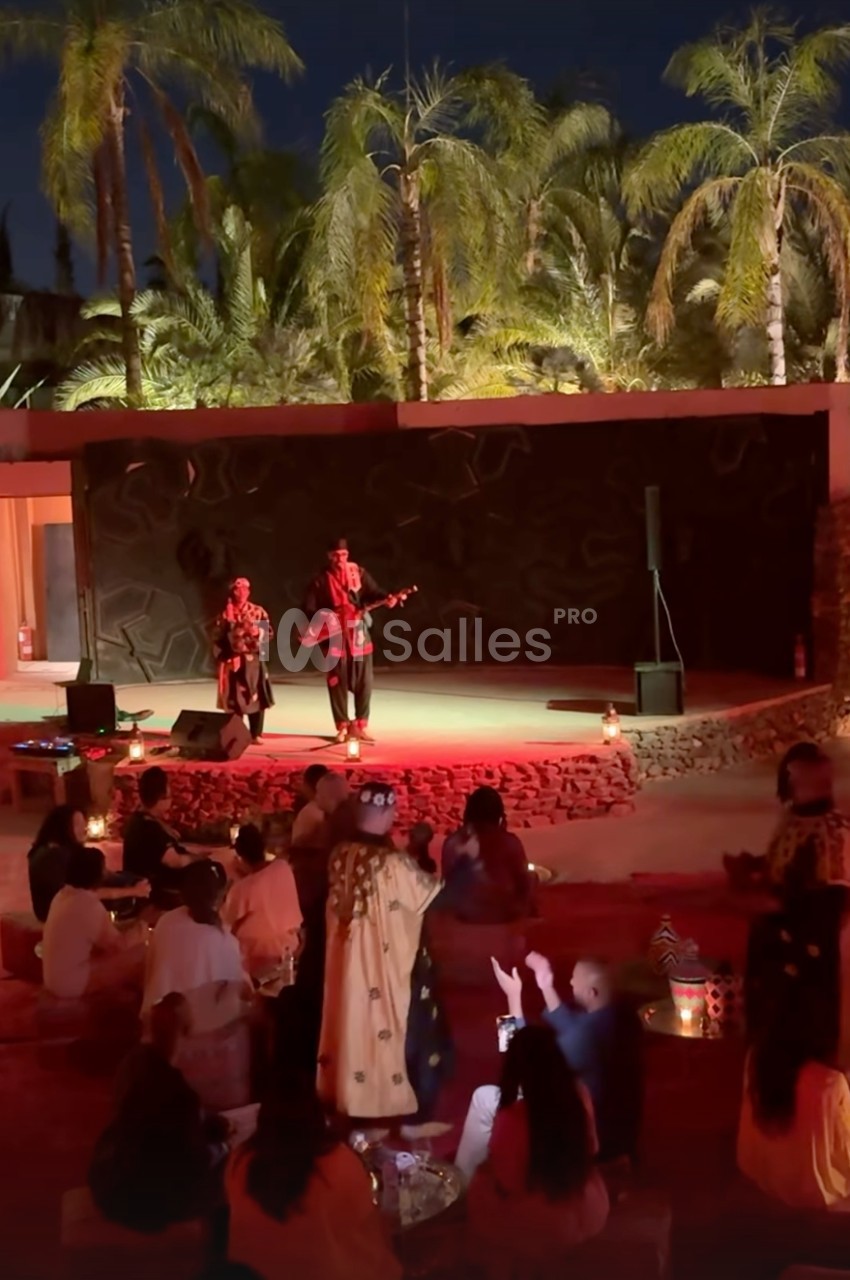 Deux musiciens jouent sur une scène extérieure éclairée, entourés de spectateurs assis sous des palmiers.