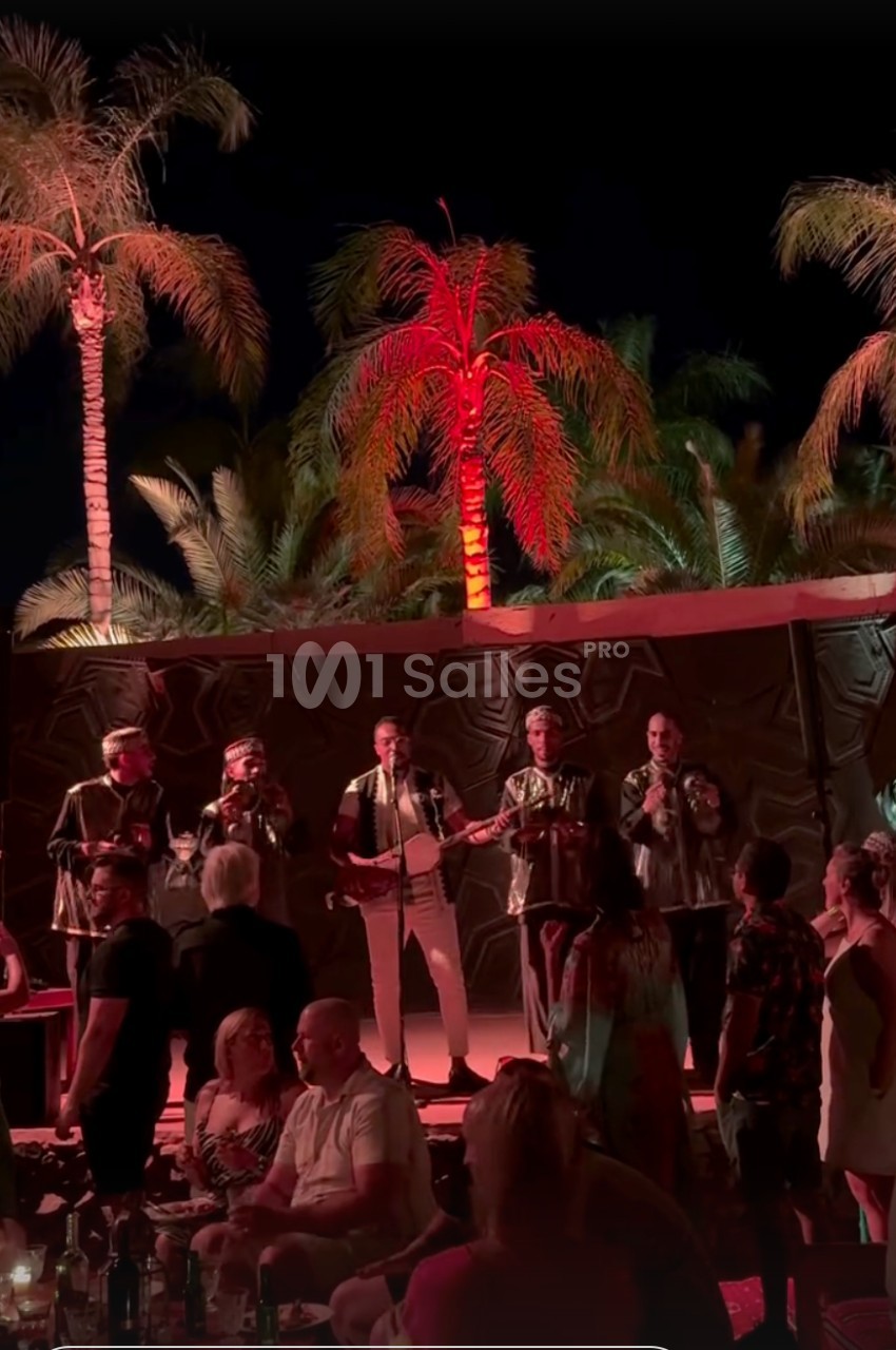 Un groupe de musiciens chante sur une scène en plein air, entouré de spectateurs et de palmiers éclairés.