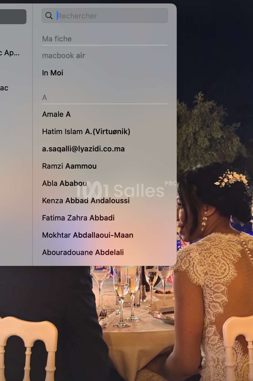 Une femme en robe de mariée assise à une table lors d'une réception en soirée, avec un écran d'ordinateur au premier plan.