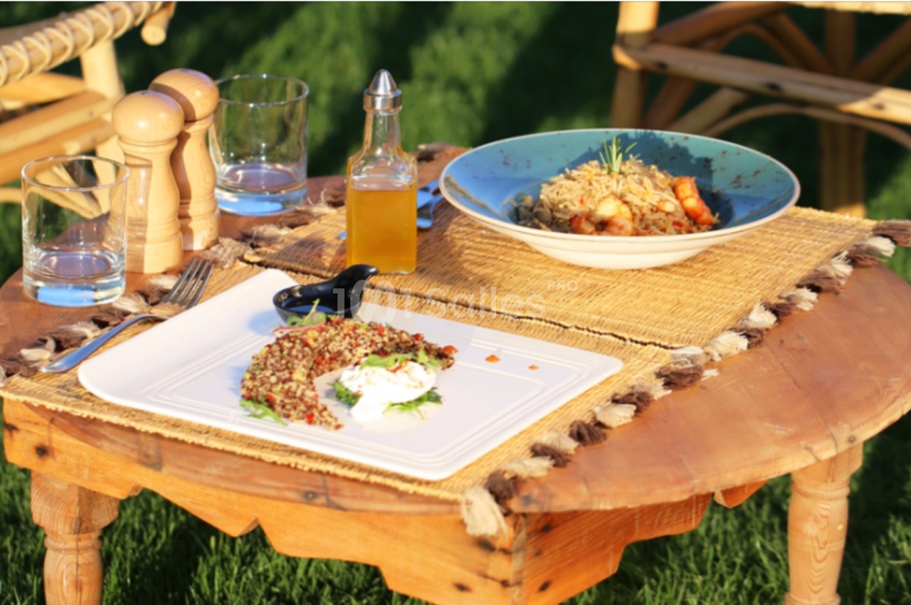 Table basse en bois sur une pelouse, avec deux plats servis, des verres, une bouteille d'huile et des moulins à épices.