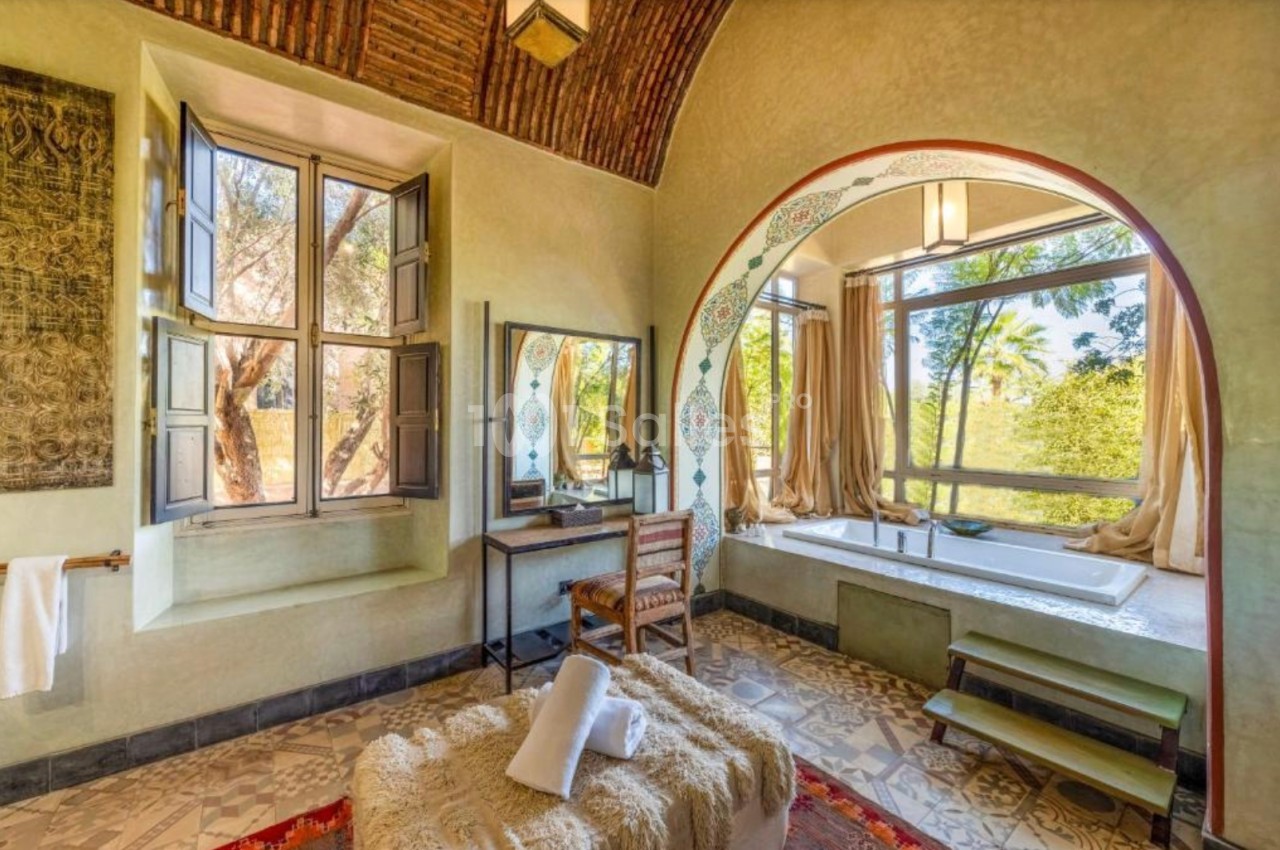 Salle de bain lumineuse avec baignoire encastrée, grande fenêtre donnant sur un jardin et décorations traditionnelles.