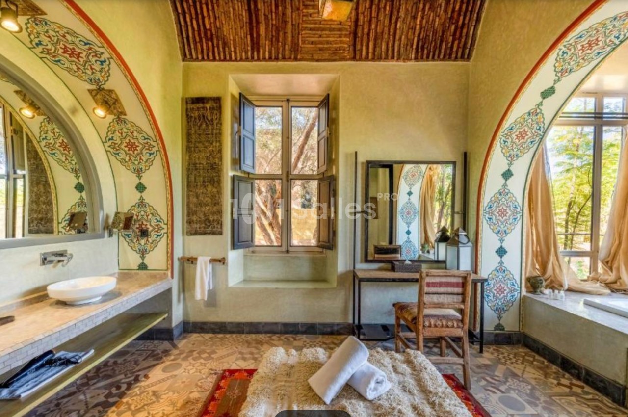 Salle de bain lumineuse avec décorations orientales, lavabo, miroir, baignoire, tapis et vue sur un jardin par une fenêtre.