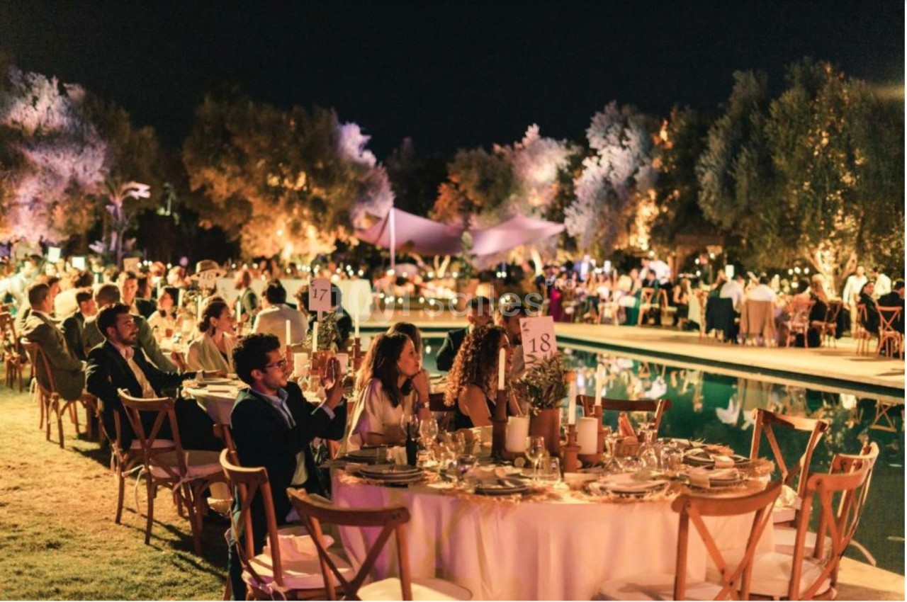 Des invités assis à des tables décorées près d'une piscine, lors d'un événement en soirée avec éclairage chaleureux.