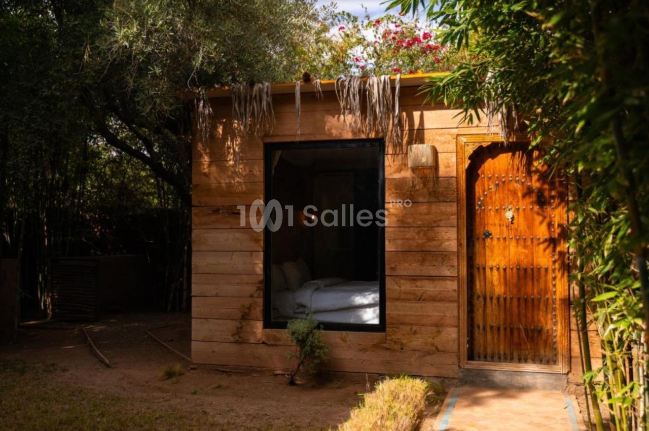 Petite cabane en bois avec une porte sculptée, une grande fenêtre et entourée de végétation.