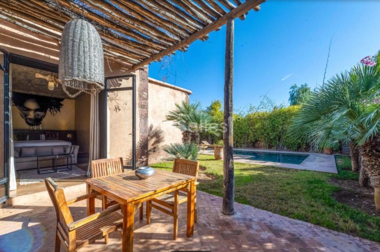 Terrasse avec table en bois, pergola en branches, vue sur jardin arboré et petite piscine sous un ciel bleu.