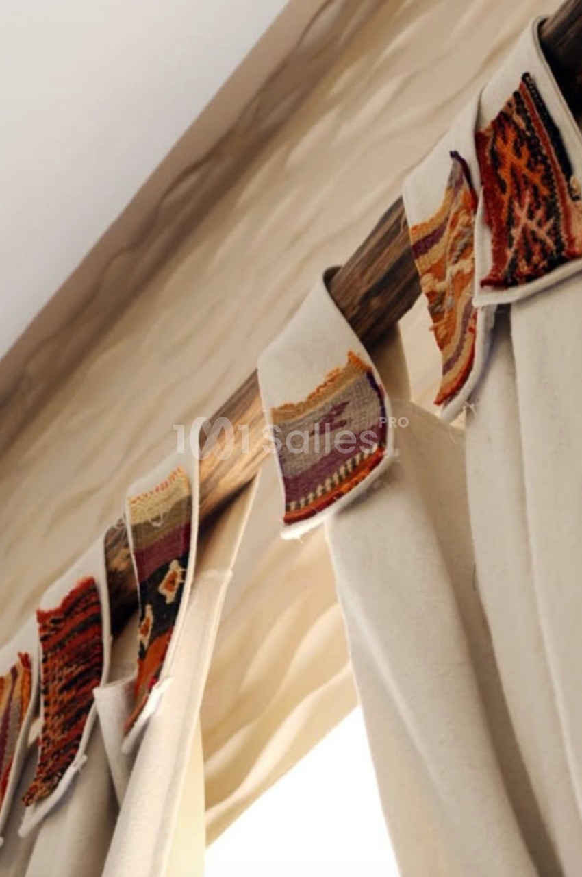 Rideau beige suspendu à une tringle en bois, décoré de bandes de tissu aux motifs colorés et géométriques.