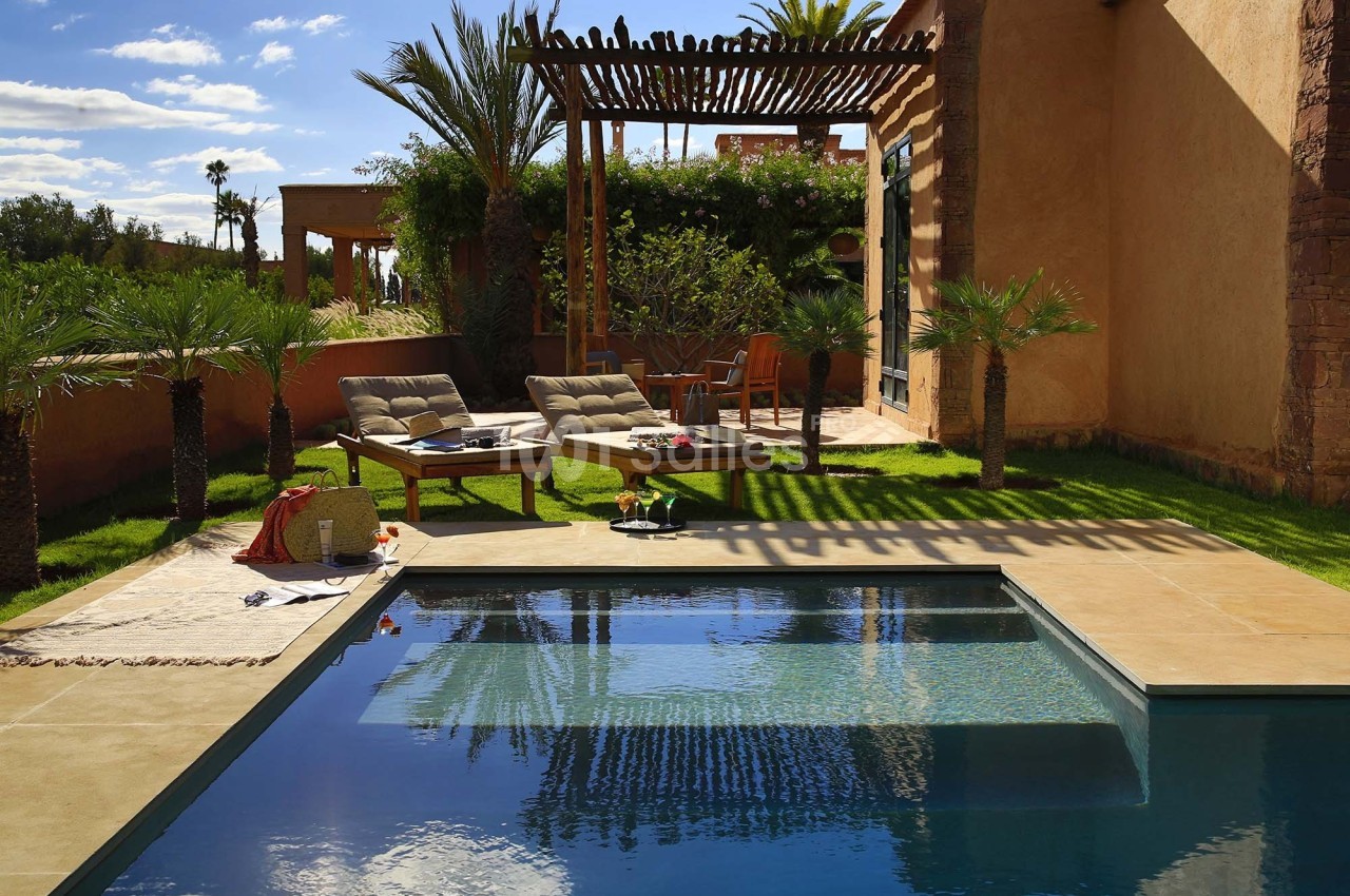 Piscine extérieure entourée de chaises longues, palmiers et pergola en bois dans un jardin ensoleillé.