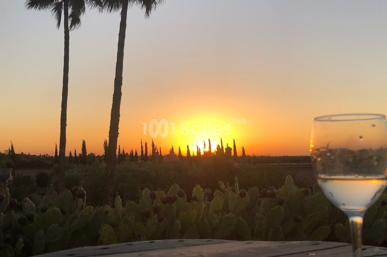 Coucher de soleil derrière des palmiers et des cactus, avec un verre de vin posé sur une table au premier plan.