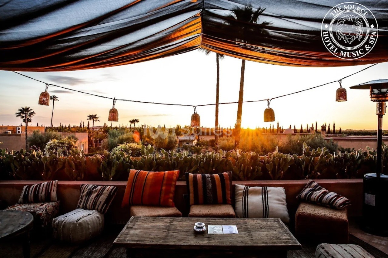 Terrasse avec mobilier en bois et coussins, vue sur un coucher de soleil, palmiers et végétation en arrière-plan.