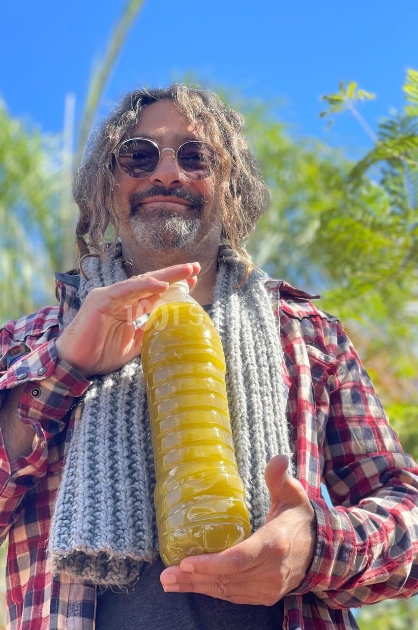 Homme souriant tenant une bouteille remplie de liquide jaune, en extérieur sous un ciel bleu avec des plantes en arrière…