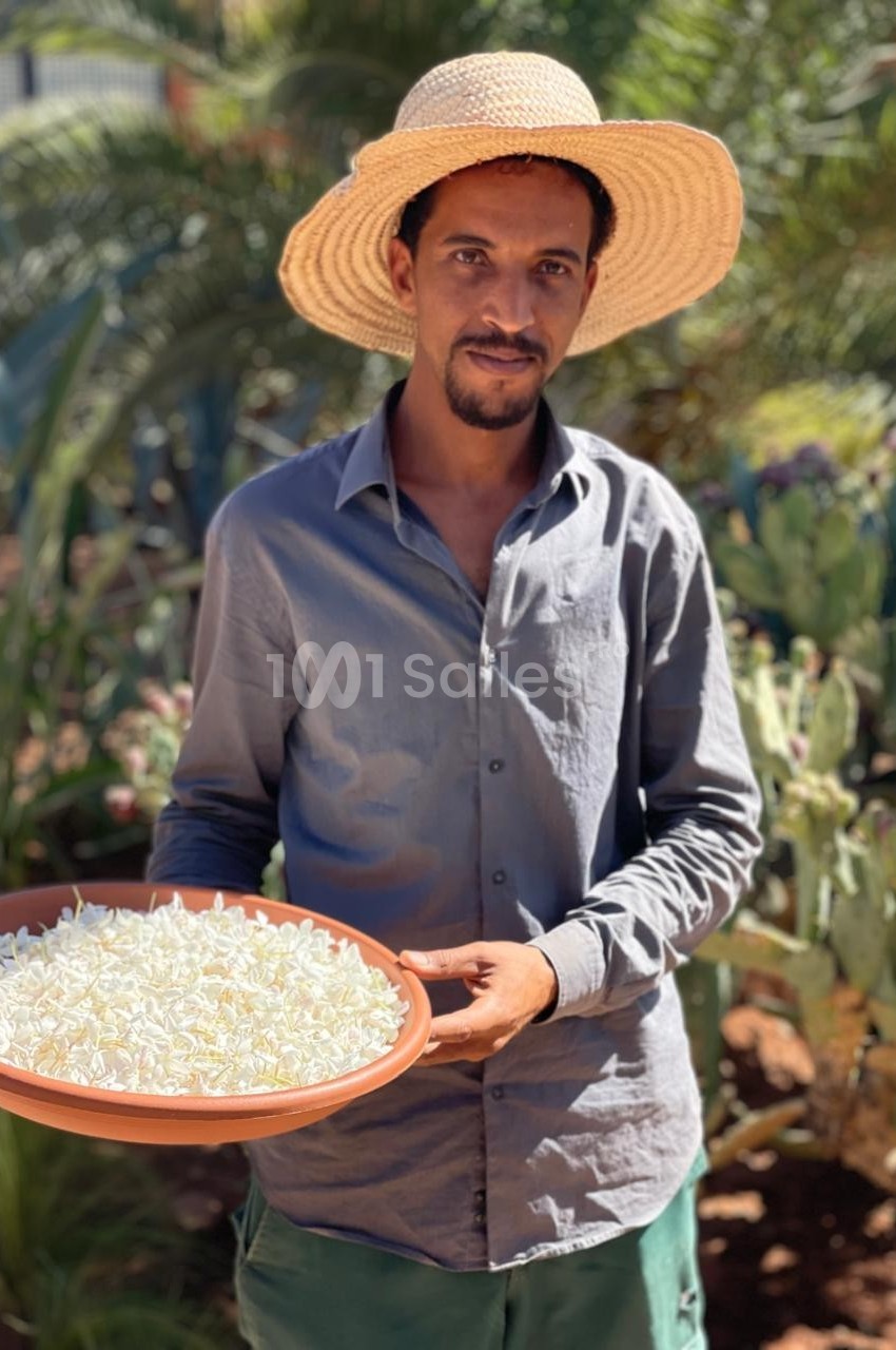 Homme portant un chapeau de paille, tenant un plateau rempli de petites fleurs blanches dans un jardin ensoleillé.