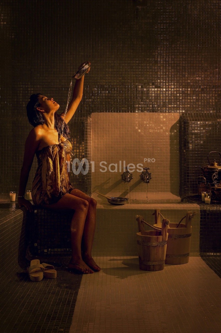 Femme assise dans un hammam, tenant un tissu humide au-dessus de sa tête, entourée de seaux en bois et bougies.