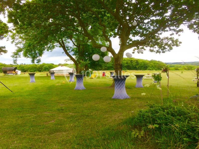Tables hautes décorées sous des arbres avec des lanternes suspendues, dans un espace vert en plein air.