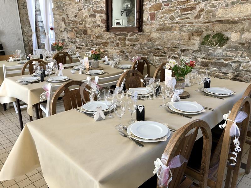 Salle de réception décorée avec des tables dressées, nappes beiges, vaisselle blanche et chaises ornées de rubans.