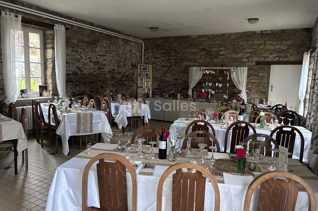 Salle de réception décorée avec des tables dressées, nappes blanches, chaises en bois et murs en pierre apparente.
