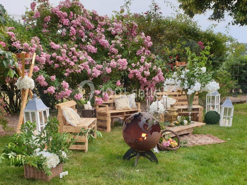 Mobilier en bois, lanternes et globe métallique décoratif dans un jardin fleuri avec des arbustes roses en arrière-plan.